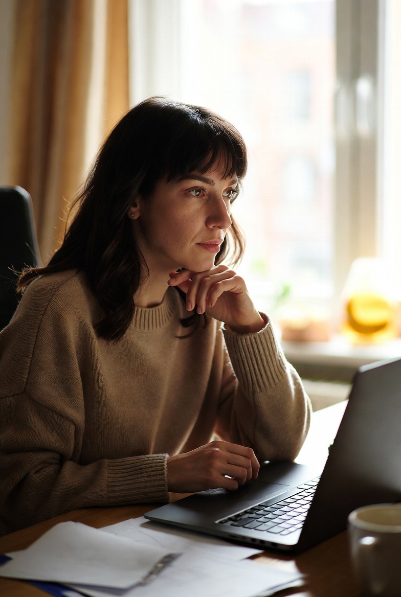 Woman in her 30s experiencing ADHD task paralysis, sitting thoughtfully at her home desk with laptop and papers, soft natural lighting, supportive atmosphere