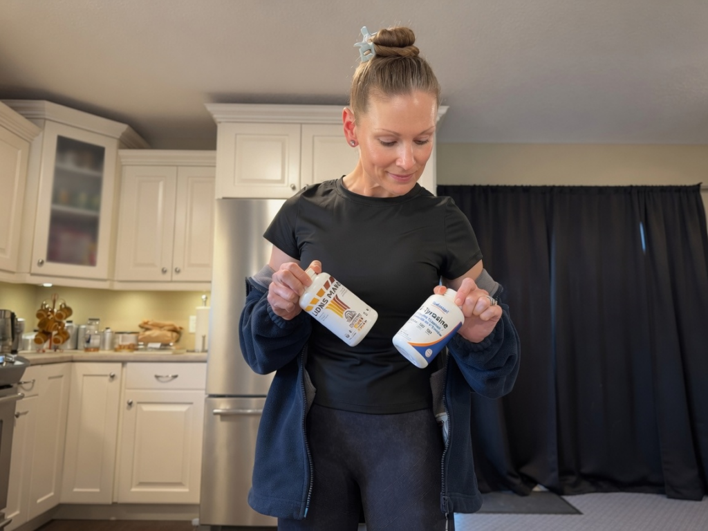 Mere stands in her kitchen holding two supplement bottles—Lion’s Mane and L-Tyrosine—while smiling softly. A personal snapshot highlighting part of her daily natural ADHD support stack.