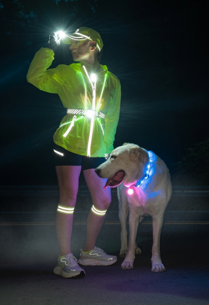 Woman in bright yellow hi-vis LED jacket with headlamp, reflective belt, and leg bands standing at night with her white dog wearing a glowing LED collar, demonstrating full safety gear for rural dawn walks and runs
