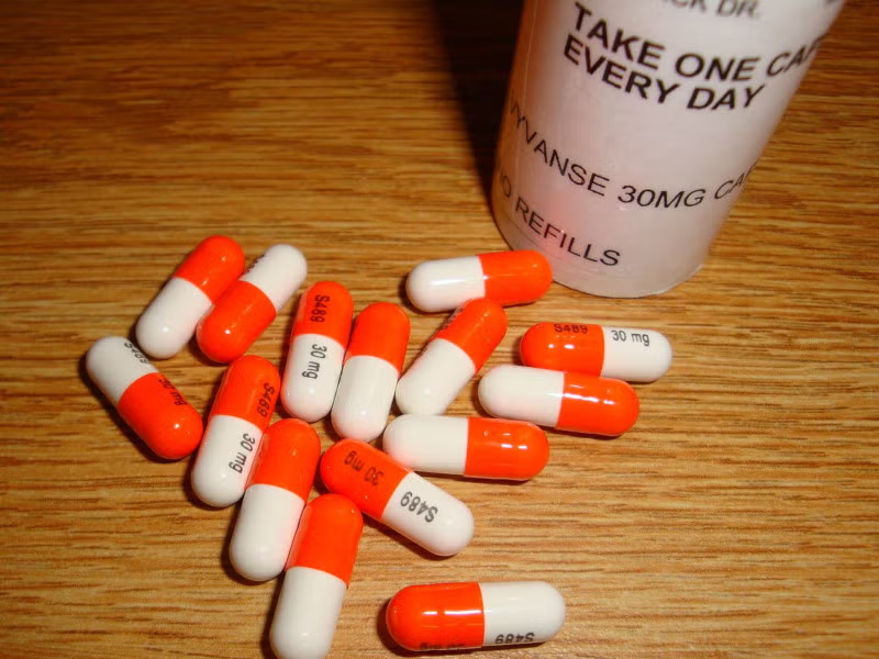 Orange and white Vyvanse 30 mg capsules scattered on a wooden surface next to the prescription bottle.